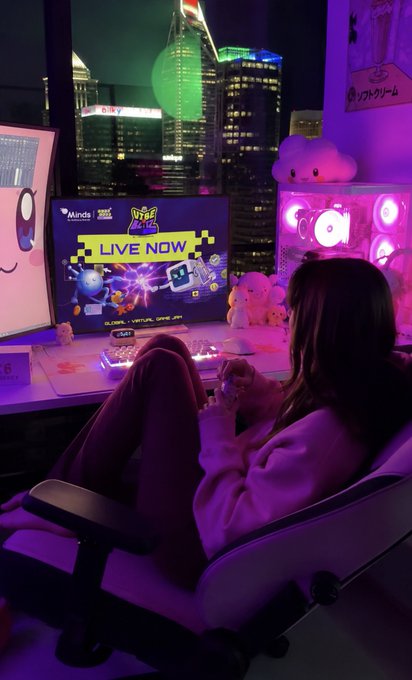 A woman relaxes in a gaming chair in a room bathed in purple and pink lighting, watching a screen displaying 'LIVE NOW' with a 'Knock on Val' and 'Minds' logo, with a city skyline visible through the window behind her.