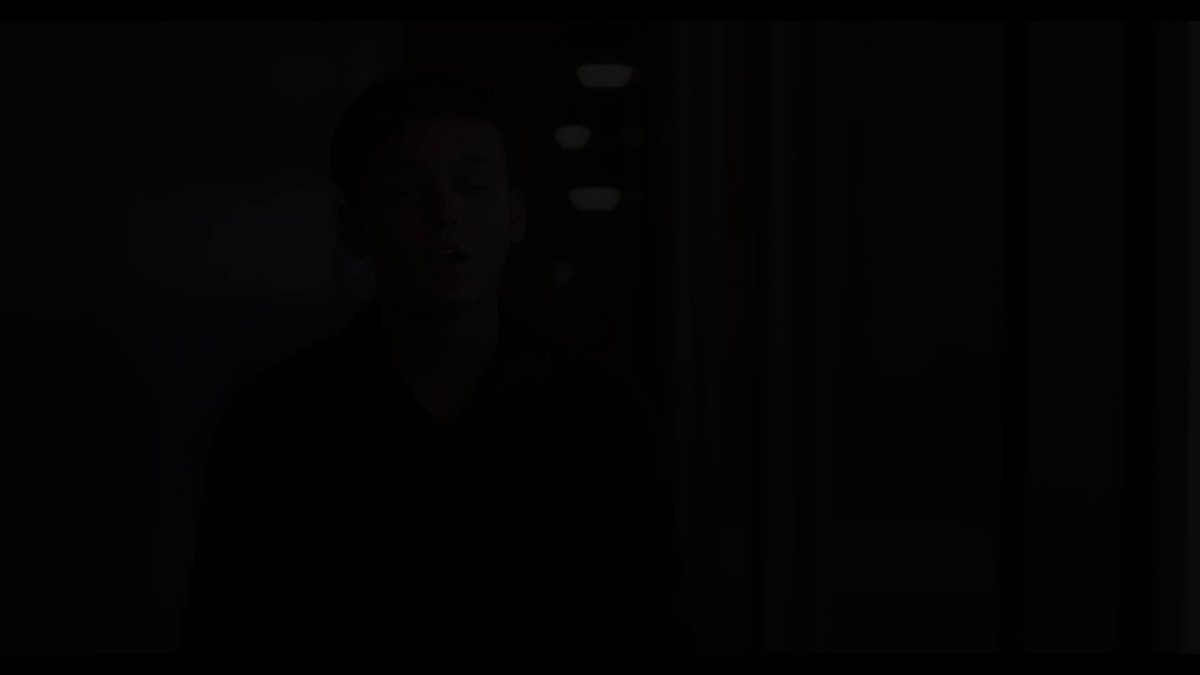 The image shows a man wearing a black shirt, standing against a dark background with his mouth open as if he's speaking or making a sound. The focus of the image seems to be on the man rather than the content displayed on the screen behind him.