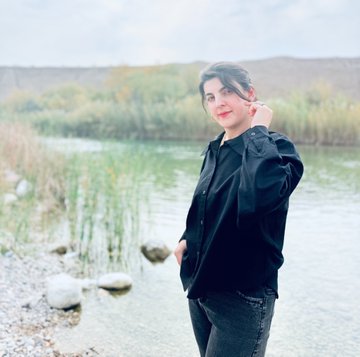 A portrait photograph shows a woman with dark hair, wearing a black jacket and dark jeans, standing by a lake with grassy hills and a cloudy sky in the background.