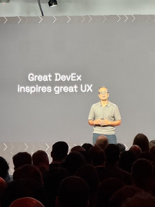 A man with glasses speaking on stage at a tech presentation, with a slide behind him reading 'Great DevEx inspires great UX' as the audience watches.