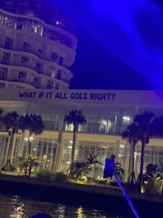 A nighttime photo shows a white building with the text 'WHAT IF IT ALL GOES RIGHT?' displayed on its facade, with palm trees and a boat visible in the foreground.