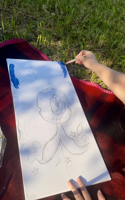 A person is painting on a canvas outdoors on a grassy area, applying blue paint to a sketch that features a central rose-like design with stars and other elements.