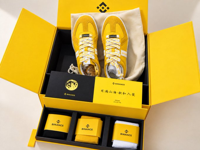 A Binance-branded merchandise box containing yellow and white sneakers, a card with Binance branding, and rolled socks featuring the Binance logo and yellow diamond symbol.