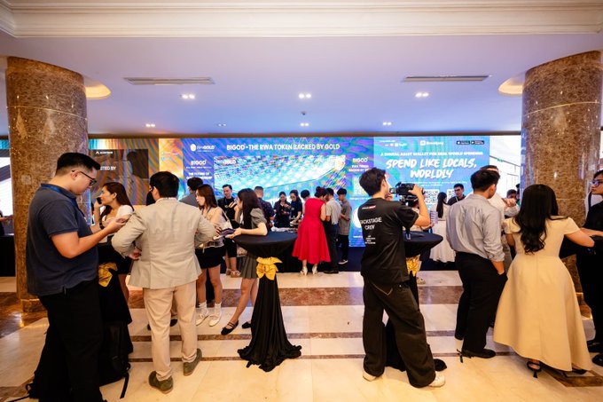 Attendees mingle at a sleek indoor event where digital displays promote "BIGOD" with the tagline "SPEND LIKE LOCALS, HOBBLY" and "BIGOD - THE BIRI TOKEN BACKED BY GOLD," aligning with the post's announcement of the BIGOD 9.9 airdrop and $2,000 reward pool for new users registering that day.