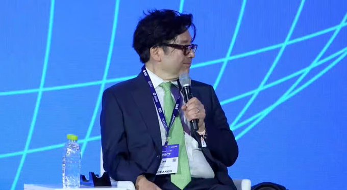A man in a suit and green tie speaks into a microphone while seated on a panel, wearing a badge that partially reads 'M AKER'.