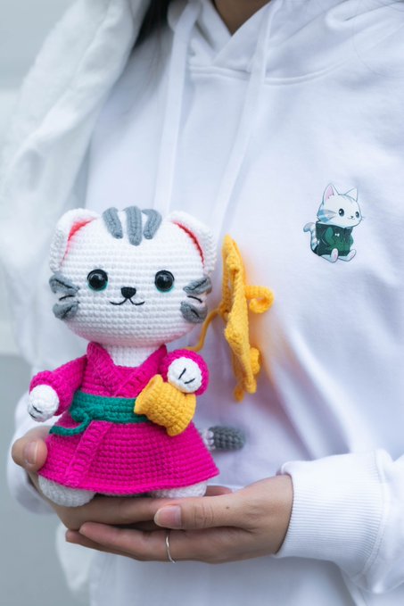 The image features a person holding up a small white stuffed animal with pink and green accents, wearing a pink robe or kimono. The stuffed animal has a yellow bell around its neck. The background of the image is blurred, but there are some text elements that read "Gmeow guys, I hosted the first Hyperliquid private event in Tokyo, Japan last year, and I'M PLANNING TO HOST ANOTHER hyper liquid / Perp DEX-themed event during @WebX_Asia this July". The person holding up the stuffed animal is wearing a white shirt.