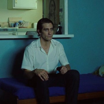 Jake Gyllenhaal sits on a purple bed in a dimly lit room with teal walls, wearing a white short-sleeved button-up shirt and dark pants, looking directly at the camera.