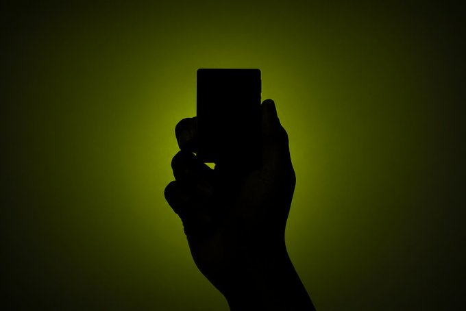 A silhouetted hand holds up a smartphone against a dark olive-green background, the device's screen emitting a faint glow from the user's fingertips. The shadowy composition evokes a sense of mystery, perfectly matching the prompt's invitation to "look closer" at the illuminated screen for hidden details.