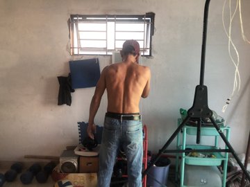 A shirtless young man in jeans stands with his back to the camera in a rustic room, looking toward a small window, with dumbbells, a fan, and exercise equipment visible around him.