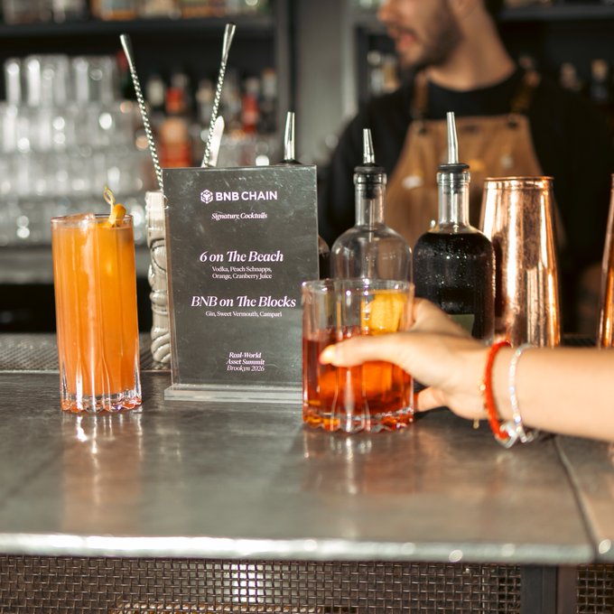 A hand reaches for an orange-hued cocktail at a bar displaying a "BNB CHAIN Signature Cocktails" menu featuring drinks like "6 on The Beach" (Vodka, Peach Schnapps, Orange, Cranberry Juice) and "BNB on The Blocks" (Gin, Sweet Vermouth, Campari), fitting perfectly with the "Shaken or stirred?" post about classic cocktail preparation.