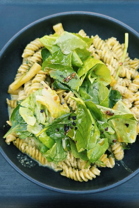 A top-down view of a black bowl containing a pasta dish, featuring spiral rotini noodles mixed with green spinach leaves, avocado slices, and red pepper flakes.