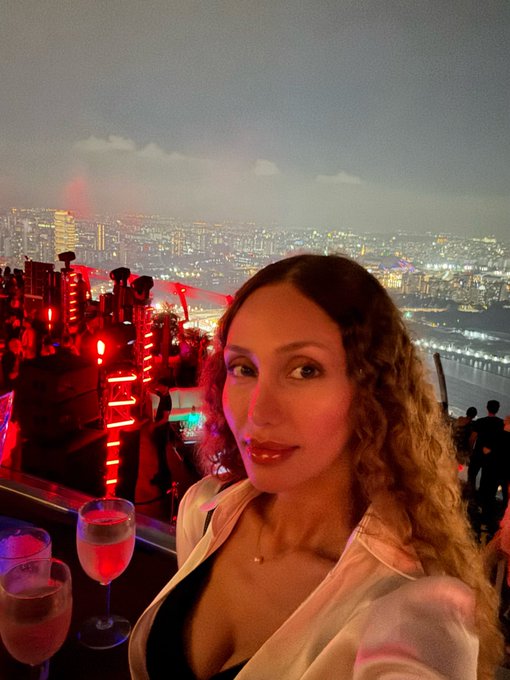 A woman with long curly hair takes a selfie at a rooftop venue at dusk, with red lights illuminating the area, wine glasses in the foreground, and a sprawling city skyline visible in the background.