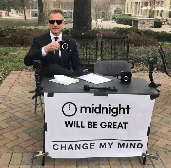 A man in a suit and sunglasses sits at a desk outdoors holding a mug, with a sign reading 'i midnight WILL BE GREAT CHANGE MY MIND' displayed on the front of the desk.