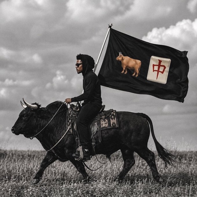 A young man in black clothing and sunglasses rides a bull across an open field, holding a black flag featuring a cow image and a mahjong tile displaying the Chinese character "中" (zhong, meaning "middle" or "center"). The bold, symbolic imagery evokes themes of virality and internet culture, visually representing the meme-driven, attention-grabbing persona associated with figures like Ansem and the $ANSEM community token.