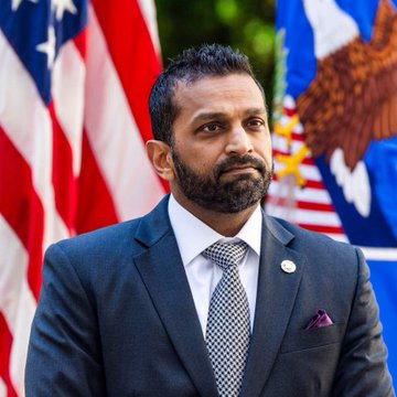 A man with a beard in a dark suit and checkered tie standing in front of American flags and a Department of Justice eagle seal.