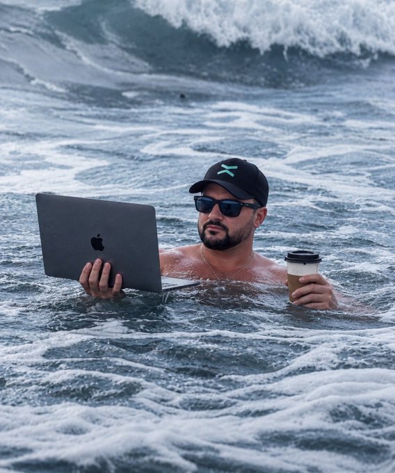 A man wearing a cap and sunglasses submerged in ocean water, holding a MacBook laptop in one hand and a coffee cup in the other.