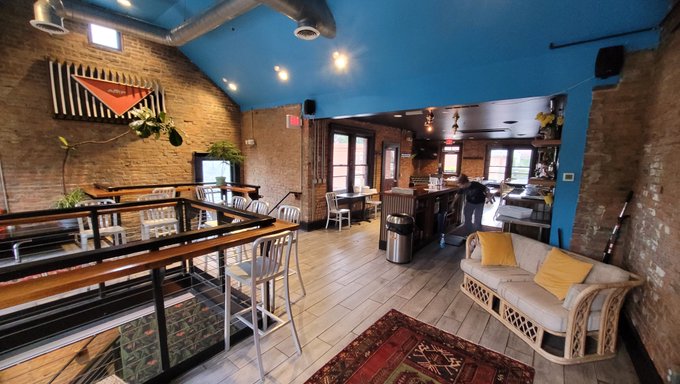 An interior view of a bar or lounge features exposed brick walls, a bright blue ceiling, a wooden bar counter with stools, and a wicker sofa with yellow pillows.