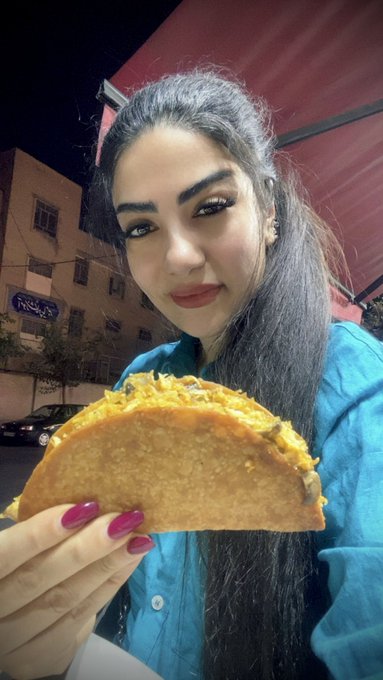 A woman with long dark hair poses at night holding a large, crispy taco, matching the "Taco Tuesday" theme of the post.