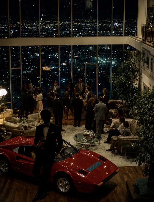 The image features a red sports car prominently displayed next to a group of people, possibly at an indoor gathering or event. The car seems to be the main attraction for this group as they stand around admiring its design and colors. The background reveals that there is a cityscape visible through the windows, which could indicate that the event takes place in an urban setting. In the post accompanying this image, there are mentions of supply chain, liquidity pools, funding, and FOMO, which further emphasize the car's significance to the gathering or event.