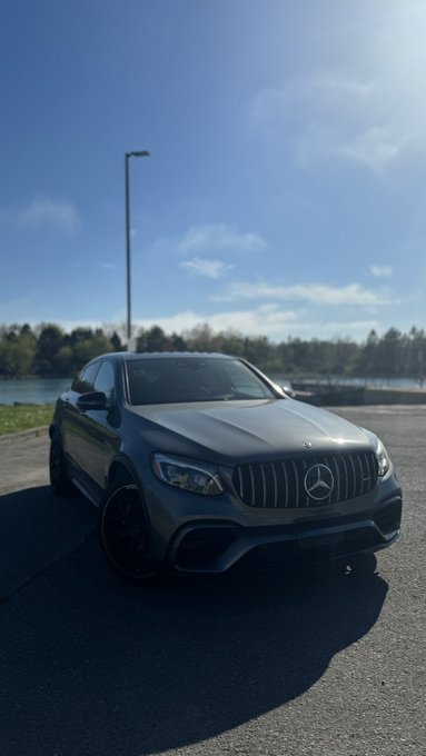 A sleek gray Mercedes-Benz (GLC Coupe, based on the styling and AMG body kit) parked by a lakeside, the iconic three-pointed star emblem prominently displayed on its grille.

The image ties to the post by playfully responding to "gm to everyone who says it back" with the brand "Mercedes" — a clever wordplay using the luxury car's name as the reply.