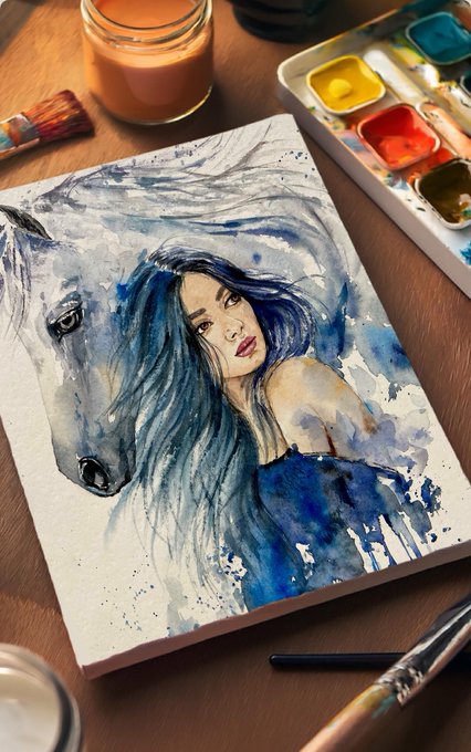 A watercolor painting on a sketchbook depicting a woman's face merged with the head of a white horse with a blue mane, surrounded by art supplies including brushes, a candle, and a paint palette.
