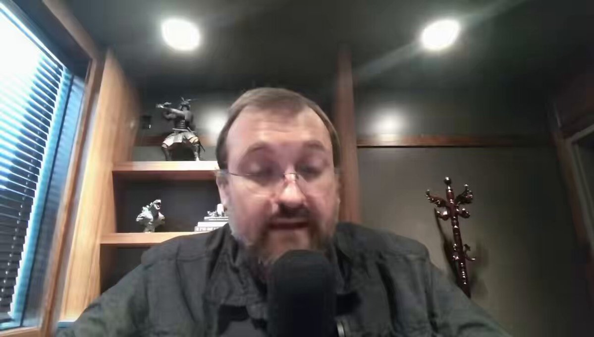 The image shows a man wearing glasses sitting at a desk with a microphone in front of him. He appears to be talking into the microphone or recording a video call. The background features a window, suggesting that he might be indoors. There are also several books on the desk and shelves nearby, indicating that this could be an office setting.
