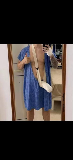 A mirror selfie showing a person in a blue hospital gown with an arm sling, taking a photo with a smartphone.