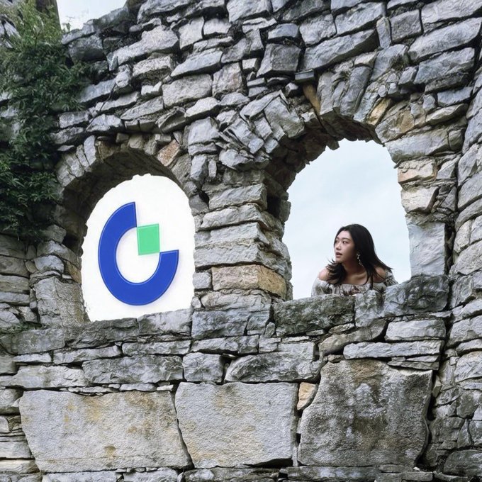 A young woman looks out from a circular window of an ancient stone wall ruin, with a stylized blue and green 'C' logo digitally placed in an adjacent window opening.