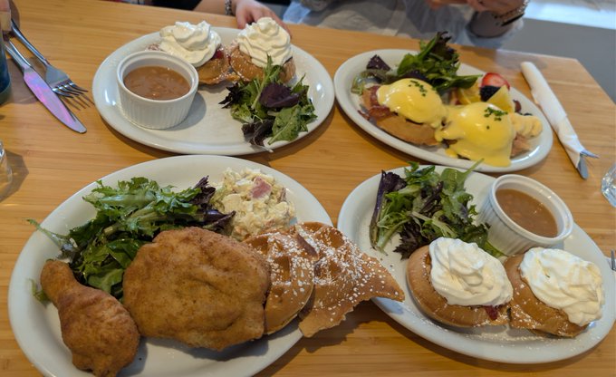 A spread of brunch dishes on a wooden table, including eggs Benedict, eggs Florentine, fried chicken with waffles, and side salads.