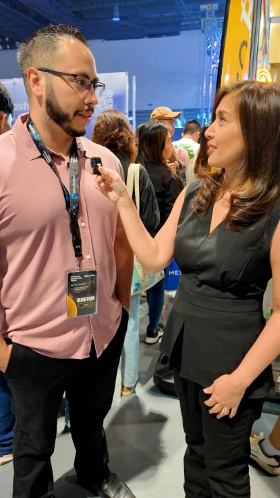 A man wearing a pink shirt and glasses, with an "INDONESIA" lanyard and "GRAND FINALIS" badge, is being interviewed by a woman in a black outfit holding a microphone at what appears to be a convention.