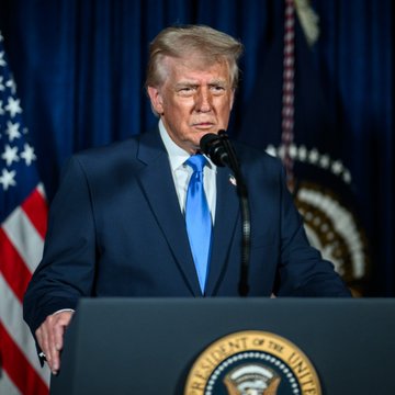 Donald Trump in a dark suit and blue tie speaking at a podium with the presidential seal, with American flags and official drapery in the background.