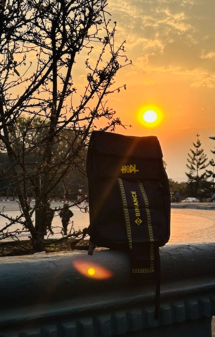 Photo of a black 'BINANCE' branded backpack resting on a railing, backlit by a bright orange sunset with tree silhouettes in the background.