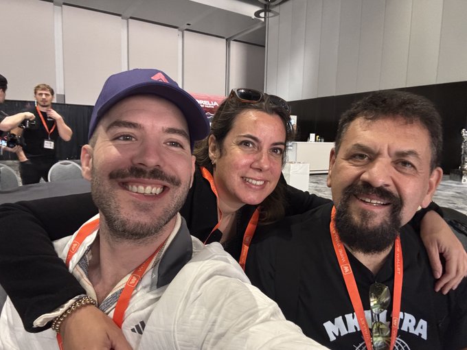 Three people smile for a selfie at what appears to be a conference, with one person wearing a 'MKULTRA' shirt and orange lanyards visible.