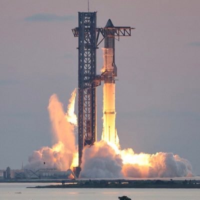 A large rocket launches from a launchpad, billowing massive clouds of smoke and bright orange fire as it ascends into a hazy sky.