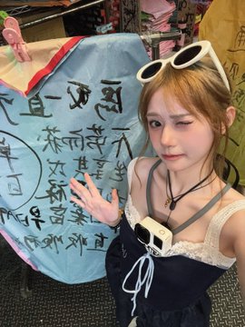 A young woman wearing white sunglasses on her head poses in a blue and black dress with a small camera strapped around her neck, standing beside Chinese calligraphy on a blue cloth.
