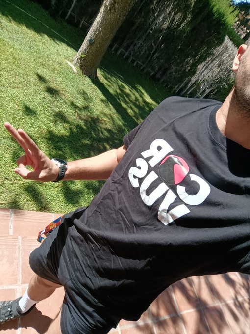 A side-profile photo of a man wearing a black t-shirt with a pink and white '1976' design, standing on a tiled patio with grass and a tree in the background.
