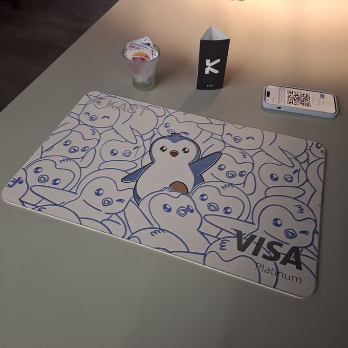 A Visa Platinum card featuring a Pudgy Penguin design is displayed on a gray surface alongside a KAST-branded item and a smartphone with a QR code. The penguin-themed card directly connects to the user's request for a Pengu card from KAST, tying the financial product to the Pudgy Penguins NFT brand.
