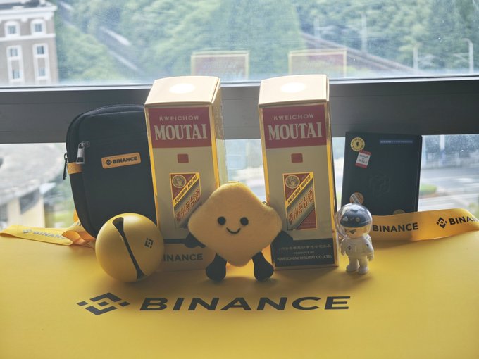 A display of Binance-branded promotional merchandise including two boxes of Kweichow Moutai liquor, Binance cases, plush toys (Binance mascot, a star character, and an astronaut), set on a yellow Binance banner.