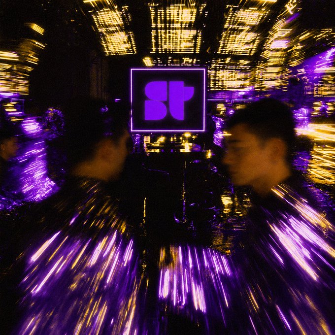 A blurred, stylized photo taken at a concert or club with two people in the foreground, featuring a purple neon sign with an 'H' logo and purple and gold lighting streaking across the frame.