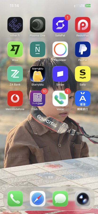 The image shows an iPhone home screen featuring a wallpaper of a young child wearing a Canon camera strap, with various financial and banking apps visible including ether.fi, SafePal, RedotPay, Wise, N26, Payoneer, PayPal, ZA Bank, Stripe, Saily, Club Sim (with 10 notifications), and HSBC, along with mobile carrier apps like Vodafone and Three. This apps arrangement relates to the post's discussion about lacking comprehensive banking options across different regions (US, Bahrain, Switzerland, Singapore) and crypto exchanges, as the displayed apps don't fully cover the international financial ecosystem described.