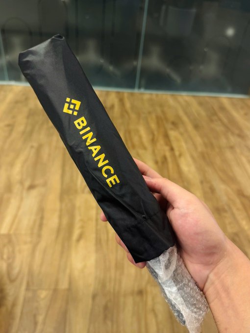 A hand holding a black umbrella with the yellow Binance logo and 'BINANCE' text, wrapped in bubble wrap.