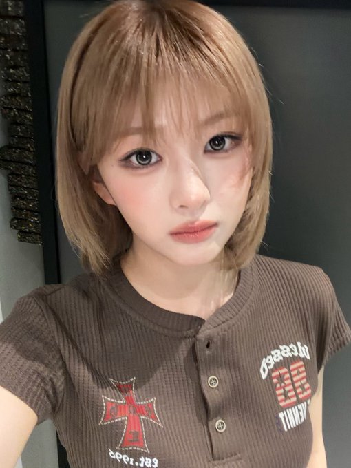 A selfie of a young woman with short blonde hair wearing a brown button-up shirt with red cross designs and text.