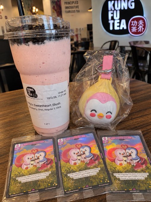 A Kung Fu Tea order featuring a 'Strawberry Sweetheart Slush' drink alongside a small pink and white plush toy and three Pokemon trading cards displayed on a wooden table.