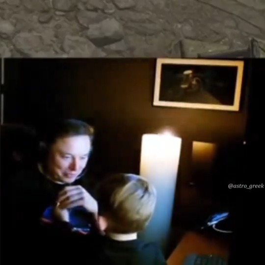 A dimly lit room showing Elon Musk wearing headphones looking at a young child with blonde hair, with the watermark '@astro_greek'.