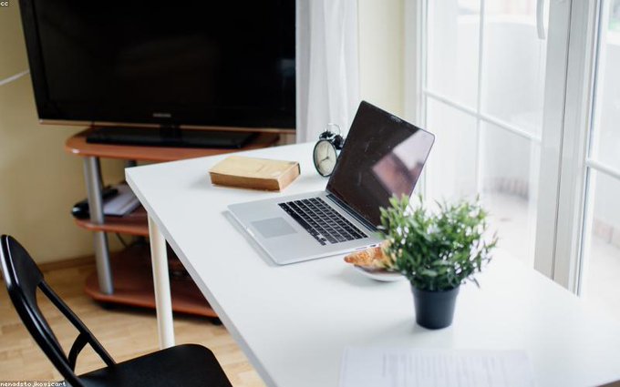 A bright home office setup with a white desk holding a laptop, an alarm clock, a small potted plant, and a TV on a stand in the background.