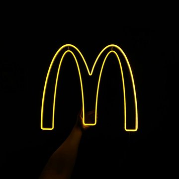 The image features a neon sign for McDonald's that reads "Mcdonald's" in yellow letters against a black background. The sign also includes an arrow pointing towards the bottom right corner, which could be interpreted as a direction or message related to the ongoing pandemic. The caption below the image states that "RUMOR: McDonald's is reportedly preparing to pilot 𝐴𝑡𝑖𝑛𝑟𝑠𝑒𝑙𝑜𝑚𝑎𝑡𝑖𝑛𝑘" and mentions the use of Bitcoin, XRP, or other digital assets.