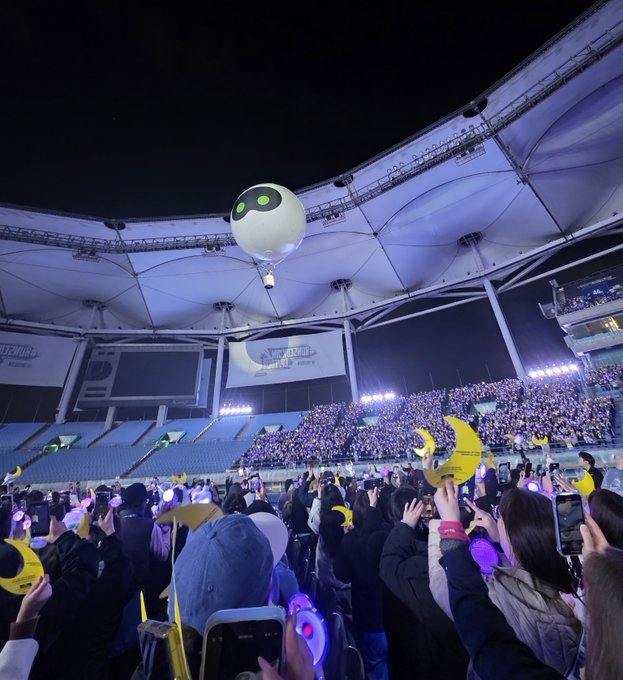 A nighttime stadium concert with a large crowd holding yellow crescent moon wands and a white balloon with a face floating in the air.