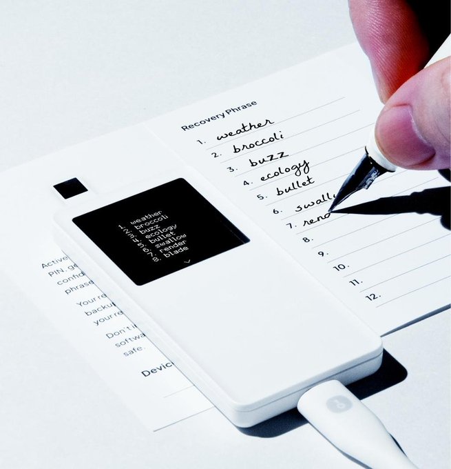 A hand uses a pen to write down a 12-word cryptocurrency recovery phrase, with the words also displayed on a hardware wallet screen.