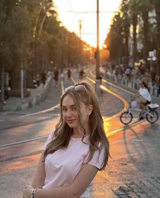 Photo of a young woman with long brown hair and sunglasses on her head, wearing a pink t-shirt and a delicate necklace, posing on a city street with tram tracks at sunset.