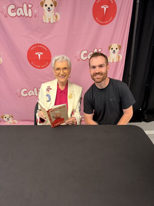 An older woman in a pink blazer holds a 'Maye Musk' book while sitting next to a young bearded man in a black t-shirt at a table, with a 'Tesla Owners Silicon Valley' pink backdrop featuring 'Cali' and a puppy logo behind them.